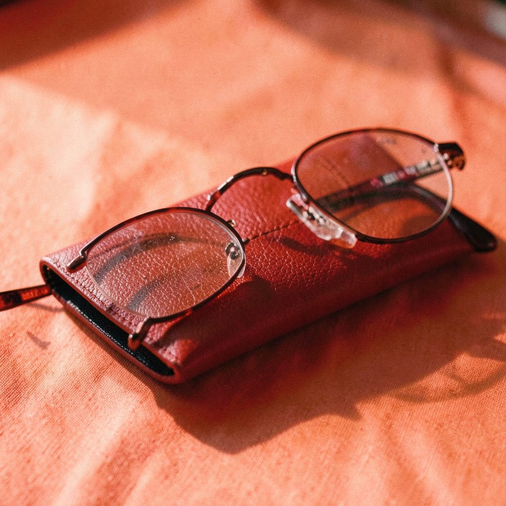 A small needlepoint eyeglass case with a delicate repeating border pattern in navy and pale gold on an ecru ground, resting on a wooden table.