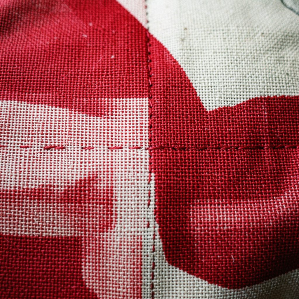 A needlepoint sampler displaying straight-stitch variations in warm gold, burgundy, and sage green threads on an even-weave fabric background.